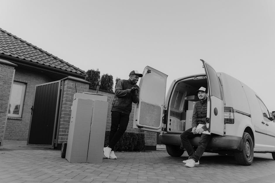 Two men engaged in home relocation activities outside a residential building during daytime, with a white van parked on the driveway. One man, standing on the pavement, is dressed in a dark jacket, trousers, and white sneakers, holding a mobile phone or small device, and appears to be inspecting or coordinating the move. The other man, sitting on the open rear door of the van, is wearing a hat, dark jacket, pants, and white footwear, and is smiling while resting. A large cardboard box and a black plastic container are positioned near the entrance of the house, indicating packed belongings ready for transportation. The van’s cargo space is visible, showing some additional boxes and possibly packaged household items. The setting includes a brick house with a tiled roof and a small garden area, and the scene is in natural light, emphasizing the loading and packing process involved in furniture transport and home relocation services. Man and Van Hillingdon, as a local removals service, handles such loading and moving operations, including packing, logistics, and transportation for house moves.