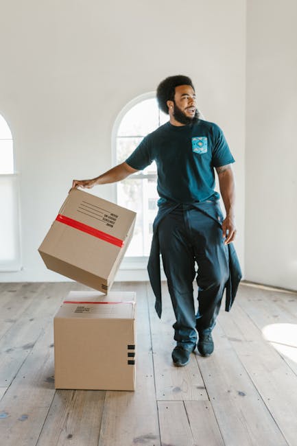 A woman with curly hair, dressed casually in a white T-shirt, blue jeans, and white sneakers, is exiting a brick building carrying a large cardboard box with smaller boxes balanced on top. She is step