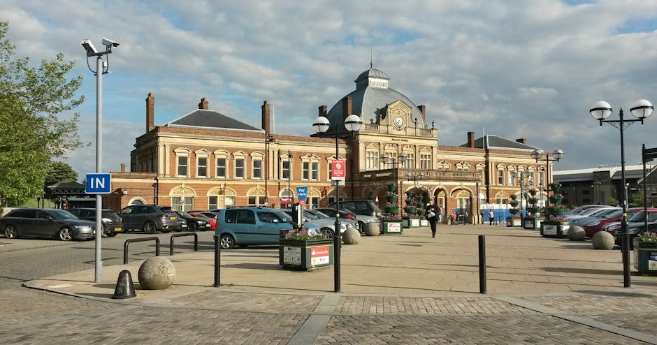 A spacious outdoor parking area in front of a historic brick building with ornate architectural details, large arched windows, and a central clock. The area is paved with concrete and bordered by black metal barriers and large spherical stone bollards. Several cars are parked along the perimeter, including a small blue van positioned near the entrance. Flower planters and tall street lamps with rounded white bulbs line the space, some wrapped with hanging flower baskets. A person is visible walking across the open paved area, which is illuminated by natural daylight with a partly cloudy sky overhead. The scene suggests a setting suitable for home relocation or furniture transport involving a moving van, with visual cues such as the open space suitable for loading or unloading goods, aligning with the house removals services described on the Brunel University student removals guide for Uxbridge, Hillingdon, provided by Man and Van Hillingdon.