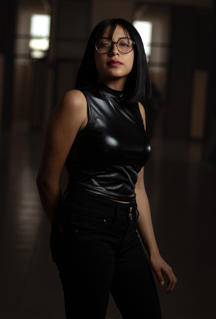 A young woman with shoulder-length black hair and fair skin stands indoors in a dimly lit corridor with a blurred background of pillars and windows. She is wearing large round glasses and a sleeveless, black, shiny leather top. Her facial expression appears neutral, and she poses with her left hand resting by her side. The lighting highlights the glossy texture of her top and her facial features, creating a subtle contrast with the darker surroundings. This image does not depict house removals or moving activities directly but could be used to represent personal preparation or organization related to home relocation or packing services by companies such as Man and Van Hillingdon.