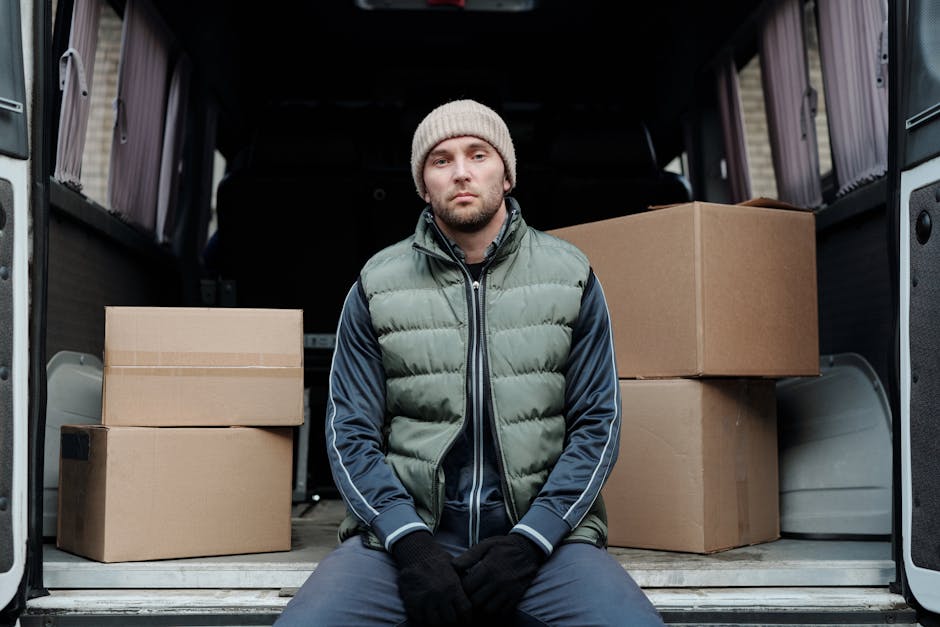 A man wearing a beige knitted beanie, a grey and navy padded vest, and dark gloves sits inside an open moving van, surrounded by several cardboard boxes of various sizes, some stacked and some placed 
