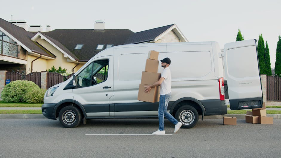 A man wearing a beige knitted beanie, a grey and navy padded vest, and dark gloves sits inside an open moving van, surrounded by several cardboard boxes of various sizes, some stacked and some placed 