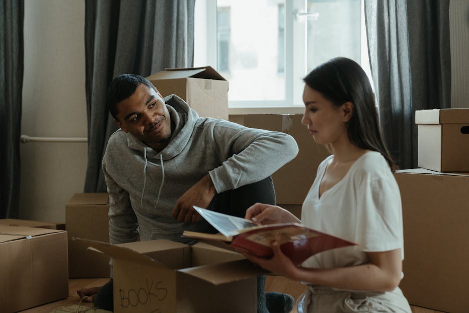 A man and a woman inside a room filled with packed cardboard boxes, some labeled with items like 'books,' during a home relocation process. The man, dressed in a grey hoodie, is sitting on the floor leaning against boxes and looking attentively at the woman, who is holding a book or folder and appears to be reading or reviewing documents. The room has a large window in the background letting in natural light, with dark curtains partially pulled back. The boxes vary in size and are stacked near the window and along the walls, some open with packing materials visible. The scene captures the logistics of furniture transport and packing in a residential setting, with cardboard and plastic wrapping materials used for protection, and a van or moving vehicle likely nearby as part of the loading process. The environment suggests an ongoing move coordinated by [COMPANY_NAME], a professional removals company, as part of a house move or furniture transport operation.