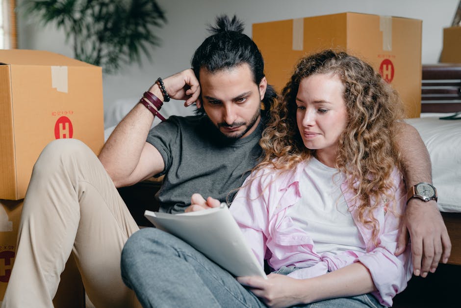 A man with long dark hair tied back and wearing a dark grey t-shirt is sitting on the floor among cardboard moving boxes, holding his head with his left hand while examining a tablet device held by a woman. The woman has long, curly light brown hair, is wearing a light pink and white striped shirt, and is looking at the tablet. They are in a room with a bed visible in the background, and the boxes around them are taped and labeled, possibly prepared for home relocation. The scene captures a moment during packing and moving preparations, with the individuals focused on planning or organizing their moving logistics. The environment is well-lit, and the setting appears to be a bedroom where house removals are underway, supporting the context of planning a professional move with companies like Man and Van Hillingdon.