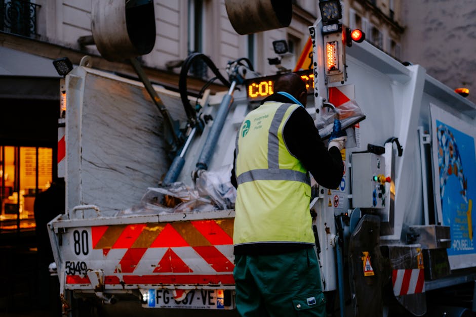 A worker wearing a high-visibility yellow vest and blue gloves operates a collection vehicle during a home removal or waste collection process in an urban setting. The vehicle features reflective red and white chevron markings on its rear and is equipped with various hydraulic and electronic controls for sorting and loading waste or recyclable materials. Inside the vehicle, visible are cardboard boxes and plastic waste, likely from packing or moving activities, arranged in the truck bed. The scene is lit with warm lighting from nearby buildings, indicating it may be early evening or late afternoon. The worker is positioned on the pavement, standing next to the vehicle and focusing on the control panel, which is partly covered with protective plastic. This image relates to furniture transport, packing, and moving logistics, relevant to home relocation services offered by Man and Van Hillingdon, with the context tied to waste removal or recycling in Hillingdon.