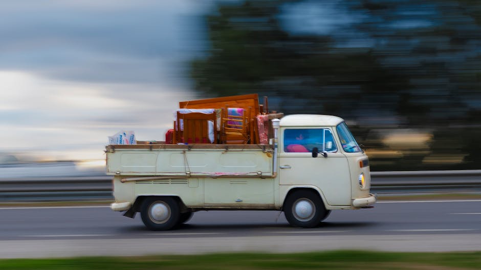 A vintage cream-colored van in motion on a paved road during daytime, with a blurred background indicating speed. The van's cargo area is loaded with various household items, including wooden chairs, cardboard boxes, plastic-wrapped packages, and small appliances, all organized to facilitate a home relocation. The furniture and boxes are secured within the cargo space, which features side panels and metal supports. Inside the vehicle, visible through the windshield and side windows, are additional items and a glimpse of the vehicle's interior. The scene reflects the loading process during a furniture transport or removal service, with the van operated by Man and Van Hillingdon, a company specialising in removals and moving logistics that efficiently handle packing, loading, and transportation tasks as part of house moving services.
