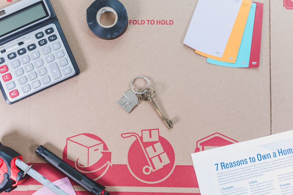 A top-down view of a cluttered workspace featuring various office supplies and moving-related items placed on a brown cardboard surface with red and white printed symbols. The objects include a calculator in the top left corner, a roll of black tape and a clear tape dispenser near the center, and a set of multi-coloured file folders in the top right. In the middle, there is a silver keyring holding a small house-shaped keychain and a brass key. To the lower left, a black marker pen lies next to a pink sticky note, and a measurement tape partially extends over a red plastic clip. A printed document titled '7 Reasons to Own a Home' is situated in the bottom right corner, overlapping the cardboard. The cardboard features red icons representing a box, a trolley, and a house, indicating themes of packing and house relocations, consistent with professional removal services. Occasionally, the workspace includes items associated with planning and logistics for home removals, aligning with the context of house moving and packing tasks.