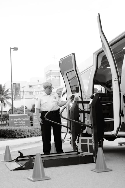 A man wearing glasses and a white shirt stands beside a small white van with its side door open, positioned on a paved outdoor area near a street. The man is holding a remote control in his right hand and observing a lifting mechanism attached to the van, which is equipped with a hydraulic lift or ramp. Several orange traffic cones surround the loading area, creating a safety perimeter. Inside the van, visible storage compartments and equipment are arranged for furniture transport or moving supplies. In the background, there are modern buildings, a palm tree, and signage indicating nearby facilities. The scene is well-lit, with natural daylight highlighting the process of loading or unloading items as part of a home relocation service, consistent with the offerings of Man and Van Hillingdon for house removals and moving logistics.