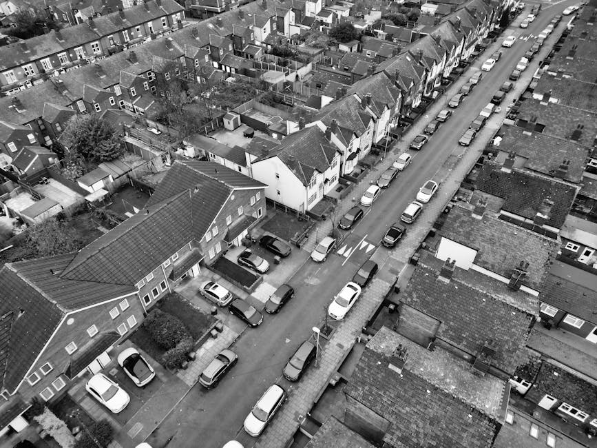 Aerial black-and-white photograph of a narrow residential street with closely spaced houses and terraced homes, lined with cars parked on both sides. The street features a central lane with parking spaces, and the pavement is flanked by houses with pitched roofs, some with small front gardens and driveways. The image captures empty spaces on the street indicating prior vehicle parking or loading areas, and the scene appears to be during daylight hours with natural lighting. The setting illustrates a typical urban or suburban neighborhood suitable for house removals and furniture transport, with visible properties awaiting or having just completed a move. Occasionally, [COMPANY_NAME] may use such visuals to demonstrate the challenges of navigating narrow access routes during home relocation or packing and moving activities.