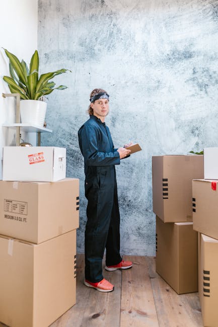 A man wearing a blue jumpsuit and a patterned headband stands indoors among several cardboard boxes, some labeled with red tape and text, in a room with a textured, light blue wall. To his left is a white shelf holding a potted plant with long green leaves. The boxes are stacked at different heights on a wooden floor, and some are open revealing packing materials. The man is holding a small clipboard and pen, appearing to be inspecting or noting details related to a home relocation or furniture transport process. The environment suggests preparation or organization for a house removal or moving service, with visible packaging and loading activities associated with professional removals by Man and Van Hillingdon.