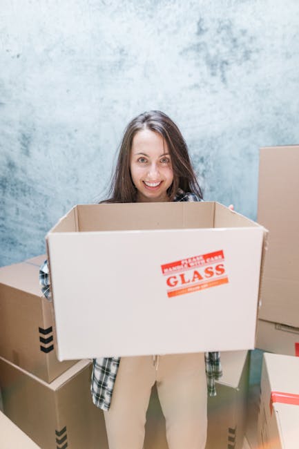 A woman with long brown hair smiling and holding an empty cardboard box labeled 'Glass' in front of her, standing indoors among several other packed cardboard boxes of varying sizes, some with black tape sealing them. The background features a blue textured wall and additional cardboard packaging, suggesting a home relocation or moving process. The scene captures the packing stage of a professional house move, consistent with services offered by Man and Van Hillingdon. The lighting is bright and even, highlighting the organized packing environment and the preparation work involved in furniture transport and moving logistics.