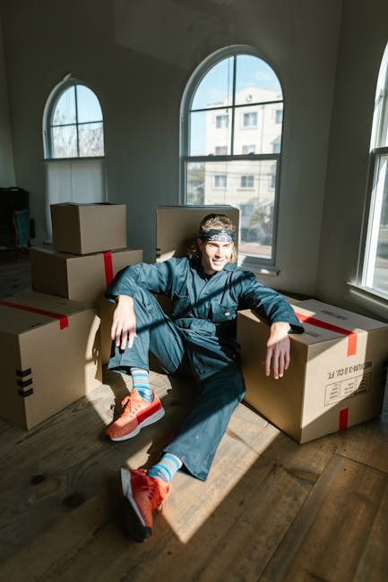 A man wearing a blue jumpsuit and a patterned headband stands indoors among several cardboard boxes, some labeled with red tape and text, in a room with a textured, light blue wall. To his left is a white shelf holding a potted plant with long green leaves. The boxes are stacked at different heights on a wooden floor, and some are open revealing packing materials. The man is holding a small clipboard and pen, appearing to be inspecting or noting details related to a home relocation or furniture transport process. The environment suggests preparation or organization for a house removal or moving service, with visible packaging and loading activities associated with professional removals by Man and Van Hillingdon.