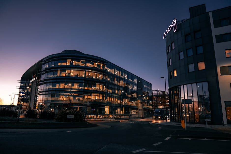 A modern office building during dusk with large glass windows reflecting interior lighting, situated on a roundabout with a small landscaped area featuring low shrubs and plants. To the right, part of another multi-storey office structure is visible, with the word 'mcy' illuminated on its upper corner. The scene includes a black van parked near the building entrance, which is used for loading furniture and packing boxes during home relocation or office removals. The pavement and road have markings, and streetlights are visible, providing illumination for the loading process. This setting exemplifies a professional move involving furniture transport and packing materials within an urban commercial environment, supported by moving services such as those offered by Man and Van Hillingdon.