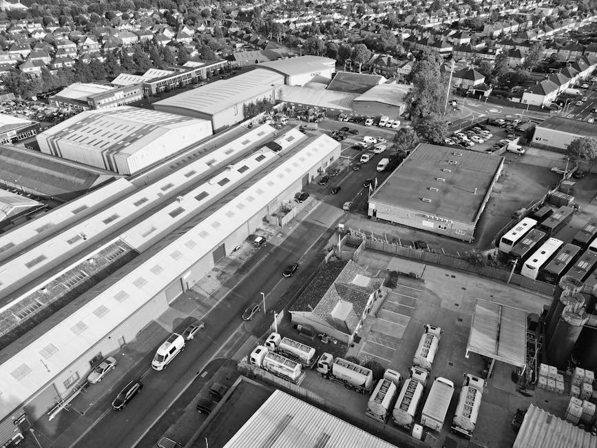 Aerial black and white photograph of a commercial and industrial area, showing several large warehouse-style buildings with flat roofs, some with skylights, and an adjacent parking lot filled with parked cars and vans. In the foreground, a man and a van from Man and Van Hillingdon are engaged in a home relocation process, with the van positioned near an entrance to one of the warehouses. Several cardboard boxes, plastic wrap, and packaging materials are visible on the pavement and inside the vehicle as part of packing and moving activities. Nearby, a trolley and lifting straps are used for loading furniture and boxes onto the van. The surrounding environment includes residential houses, trees, and a network of roads, with some vehicles actively driving along the streets. The scene captures the logistics and unloading process typical of furniture transport and home removal services, highlighting professional moving operations in an urban setting.