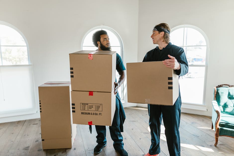Two men engaged in a home relocation process inside a well-lit room with white walls and large arched windows. One man, with dark curly hair and a beard, holds two stacked cardboard boxes secured with red tape, standing beside a third box on the floor, which is marked as medium capacity. The other man, wearing a black headband and casual dark clothing, is holding a single cardboard box while gesturing with his hand as if communicating. The room contains a green upholstered armchair with wooden accents on the right side, and the floor is made of light-colored wooden planks. In the background, there are no other objects or furniture visible, and sunlight streams through the windows, creating a bright environment suitable for packing and furniture transport during a house move. The scene reflects a loading process involving packing materials like boxes, positioning, and coordinated effort typical of professional moving services, such as those provided by Man and Van Hillingdon, with a focus on efficient relocation logistics.