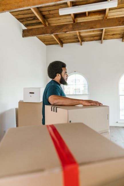 A man with dark curly hair and a beard, dressed in a blue shirt, is inside a spacious room with a high wooden-beamed ceiling and white walls. He is standing behind several large cardboard moving boxes, one of which is taped with red packing tape, positioned on a flat surface, possibly a table or the floor. The boxes vary in size, with some placed on top of each other. The room features two arched windows allowing natural light to illuminate the space, revealing an urban exterior view. The setting appears to be during a home relocation or packing process, with the man preparing the boxes for transport or loading. Visible in the background are more boxes and possibly protective packing materials, indicating an organized packing and moving operation by [COMPANY_NAME]. The scene captures a moment of preparatory furniture transport or house removal within a professional moving service context, reflecting careful packing and handling for house removals in the Hillingdon area.