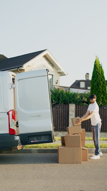 A man and a woman are standing beside an open removal van, which is loaded with numerous medium-sized cardboard boxes stacked inside. The man, wearing a black cap, white t-shirt, and earphones, is hol